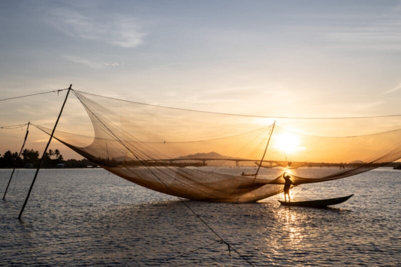 A person stands on a small boat beneath large fishing nets on a calm body of water at sunset, with the sky glowing orange and silhouettes of distant trees and mountains in the background.