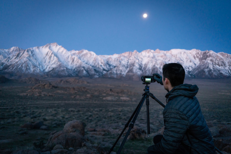 A man pointing a camera on a tripod at a snowy mountain range with the moon in the sky