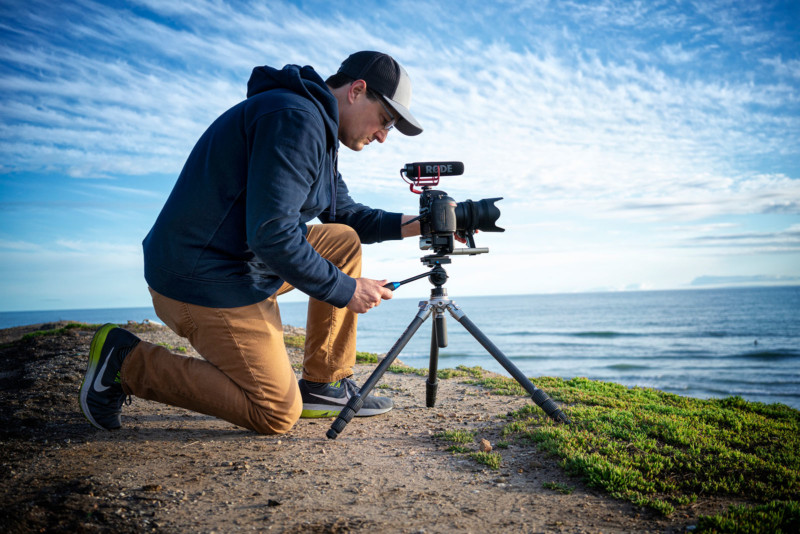 A man setting up a camera and tripod on a hill overlooking the ocean