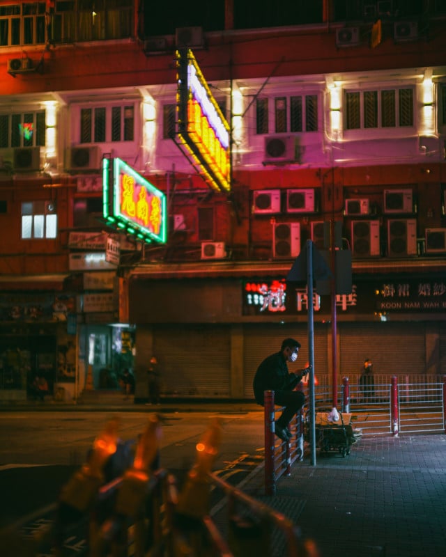 A nighttime street scene with a man sitting on a railing using his phone