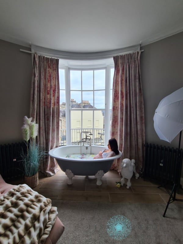 A pregnant mother sits in a milk bath next to a high window