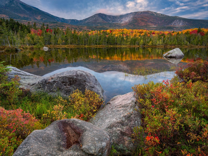 A serene mountain scene with calm water reflecting the vibrant autumn foliage of green, yellow, and orange. Large rocks and colorful shrubs are in the foreground, with a forest and majestic mountains under a partially cloudy blue sky in the background.