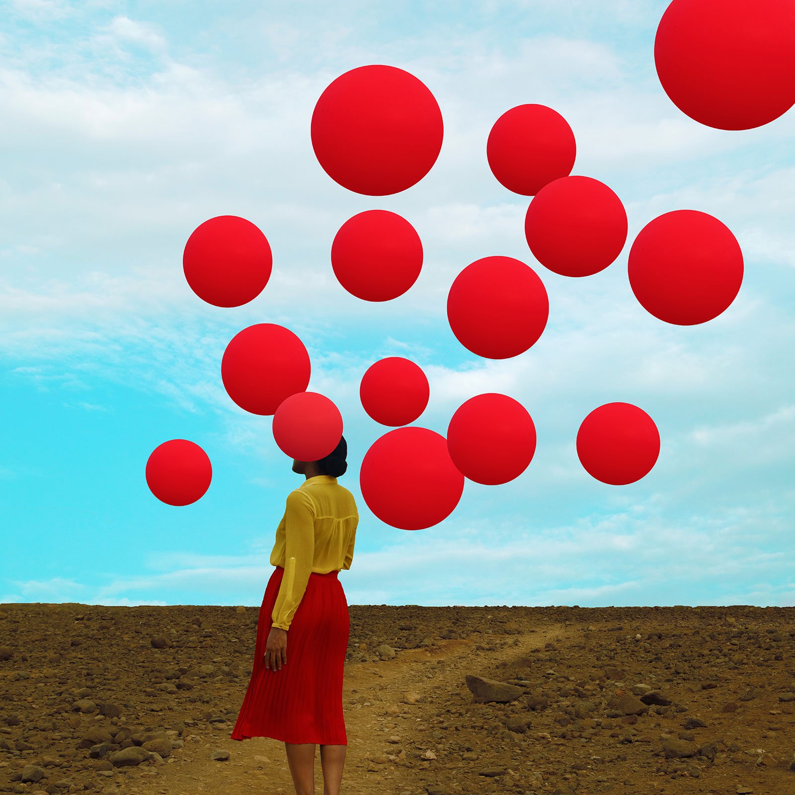 A woman in a vast brown landscape with bright red balloons covering her face and flying over her