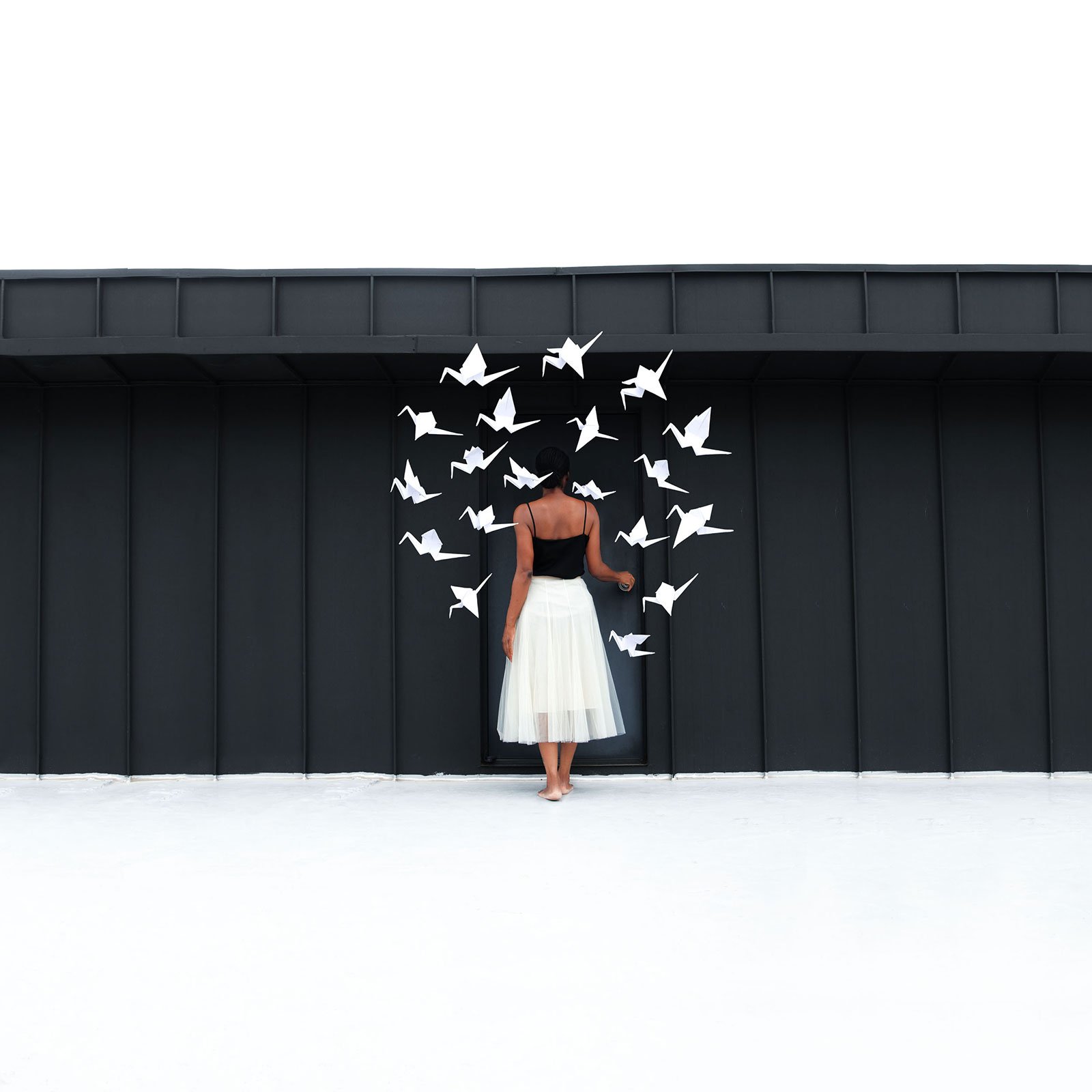 woman with white skirt and black top facing a black wooden background with paper cranes floating in a circle above