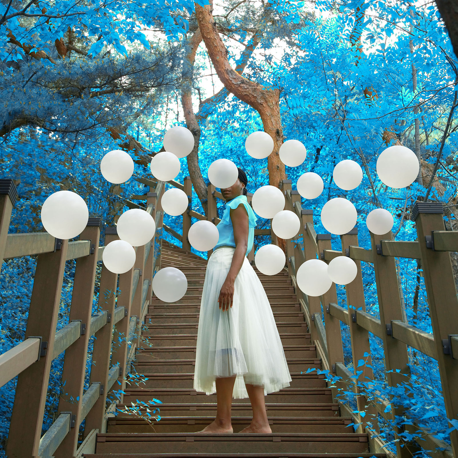 woman going up stairs, towards a bright blue forest and white balloons cover her face
