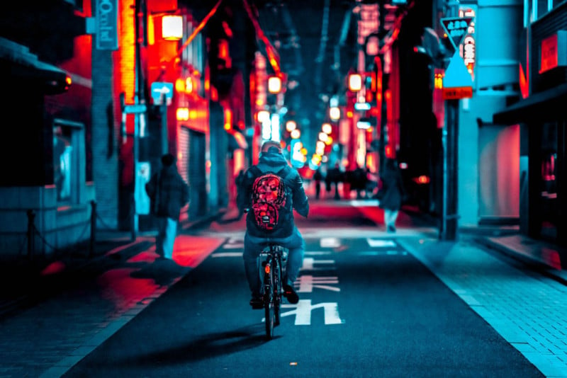 Street shot of Kyoto at night: young man riding a bicycle, riding away from the camera, he is wearing a bright red back pack and grey and blue hoodie