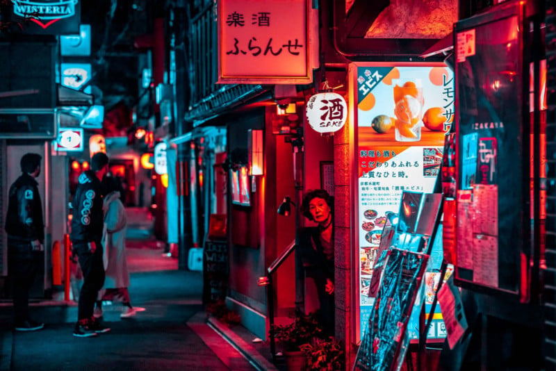 Street shot of Kyoto at night: woman peaking at street from store with multiple small shops in the distance 