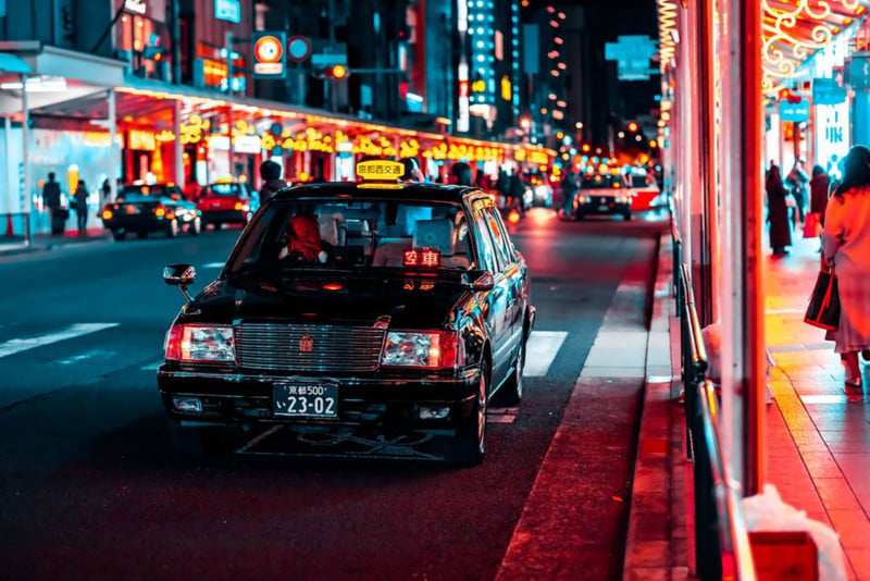 Street shot of Kyoto at night: black vehicle parked with middle aged man inside and bright city lights to his left