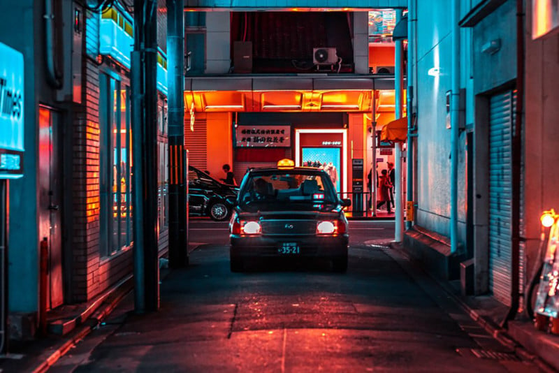 Street shot of Kyoto at night: black vehicle in alleyway, with neon lights behind. The cars headlights are on