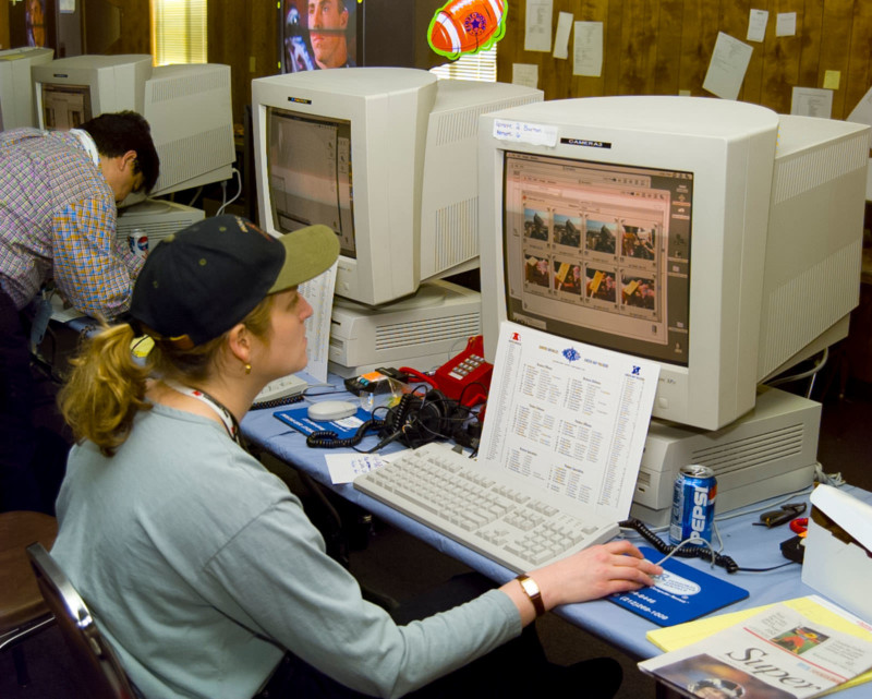 A blonde woman wearing a ballcap sits in front of a large computer workstation with a page of notes and a can of soda on the desk