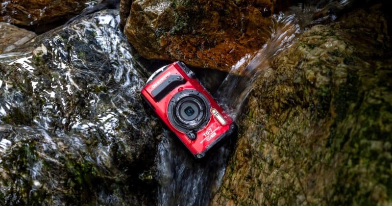 A red waterproof camera lies partially submerged in a small stream, surrounded by moss-covered rocks. The water flows gently around the camera, highlighting its rugged design.