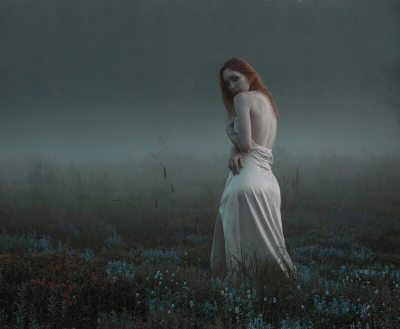 pale woman, with red hair, and a white dress, looking slightly at the camera, standing in a misty field of wild flowers