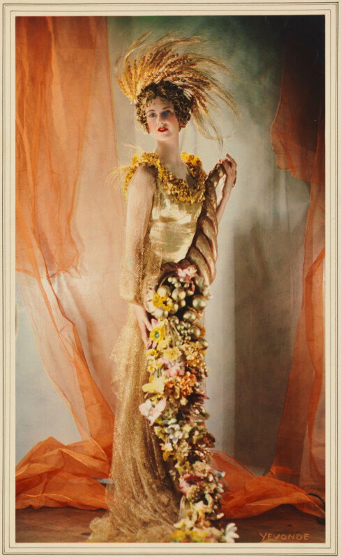 Lady Dorothy Warrender se tient dans une robe dorée avec un bouquet de printemps commercial.  Elle porte une coiffe de plumes orange qui ressemble à des flammes léchant le sommet de sa tête.