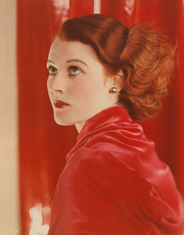 An all-red portrait of Joan Maude in close up. Her hair is red and waved, she wears a red shirt and stands against a red backdrop, looking skywards.