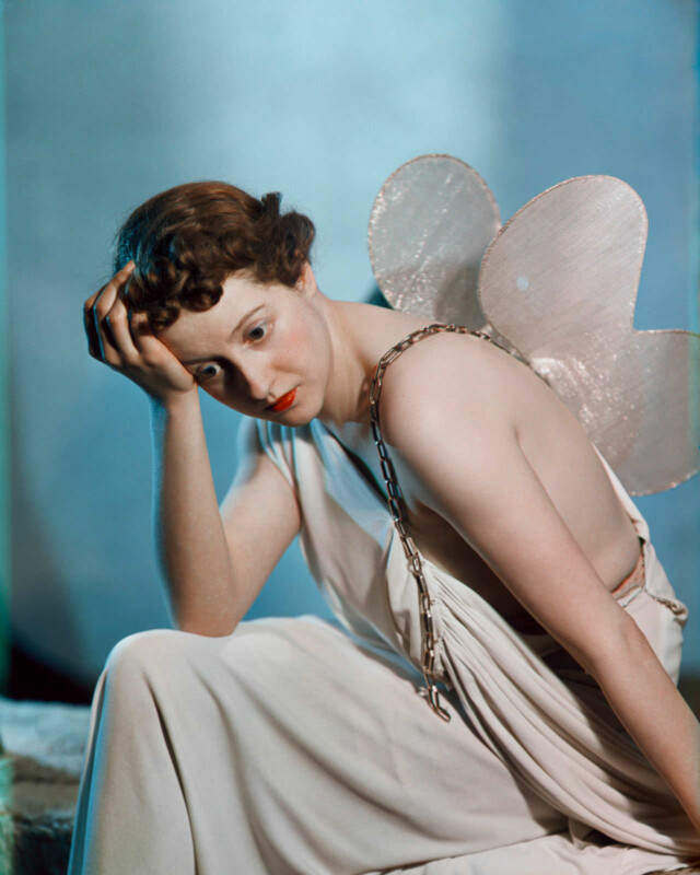 Dorothy Gisborne with her head in her hand wears a white robe dress and fairy wings, against a blue backdrop.