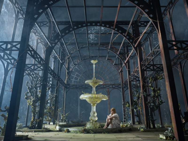 a woman sitting by a glass fountain in a large abandoned brass and blue tinted greenhouse 