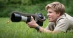 A smiling person lies on grass, holding a camera with a large lens while a snake is slithering over the lens; trees blur in the background.