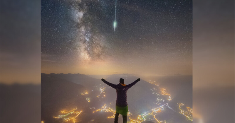 A person stands on top of a mountain as a meteor and the Milky Way Galaxy shines above them. 