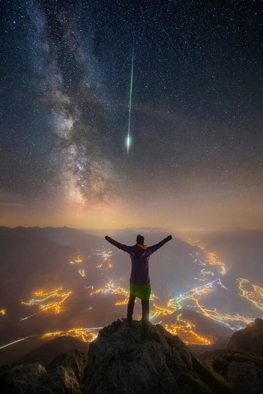 A person stands on top of a mountain as a meteor and the Milky Way Galaxy shines above them. 