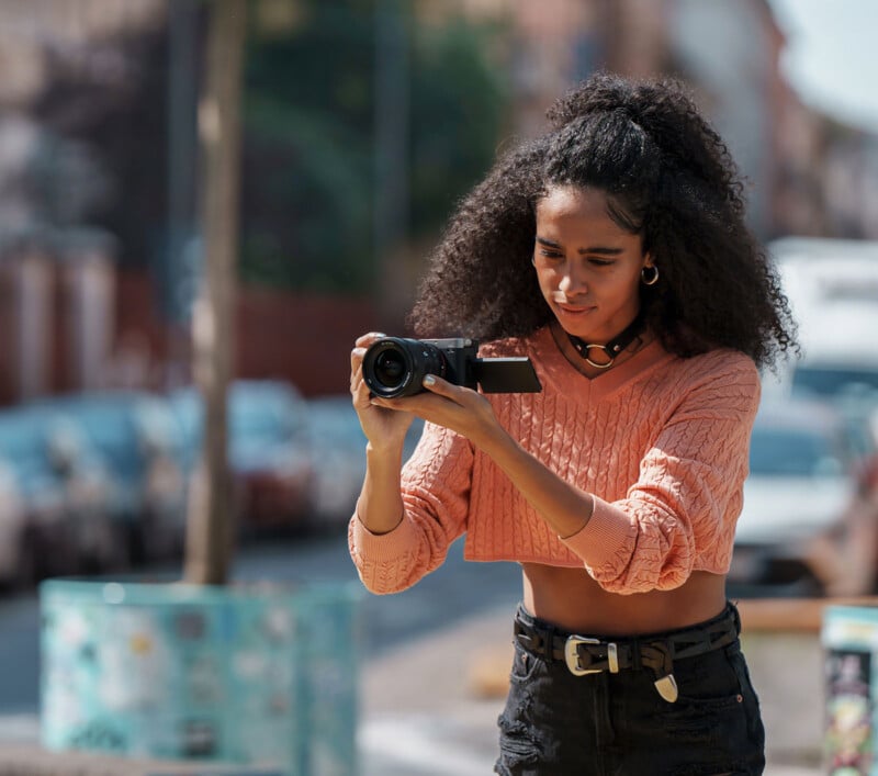 A woman with curly hair wearing a peach cropped sweater and black jeans films with a camera outdoors on a city street during the day.