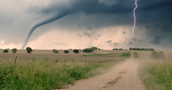 A car drives on a dirt road while a tornado and lightning are seen in the background.