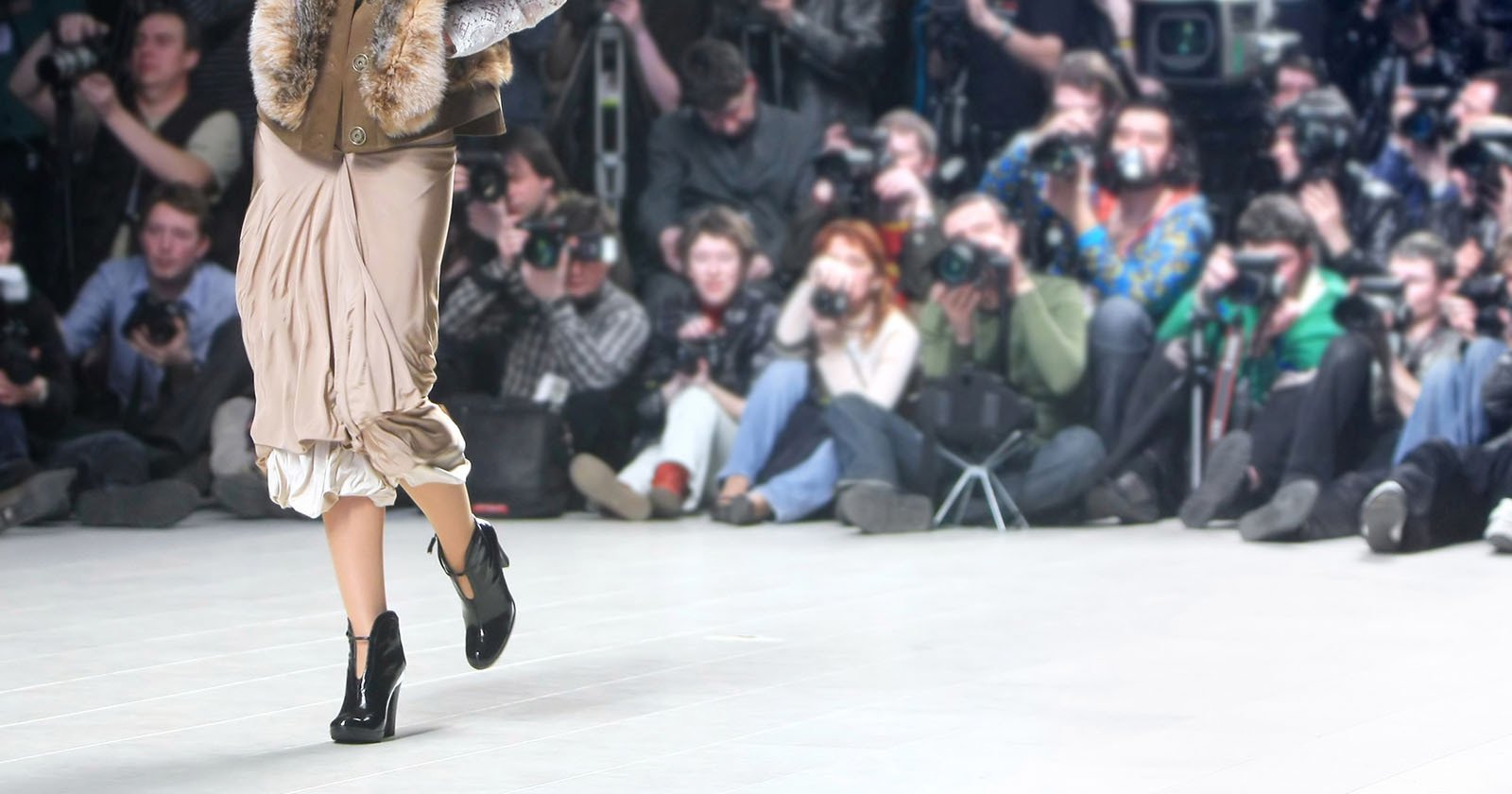 A model walking a runway is seen from the waist down and an audience and photographers are viewed in the background.