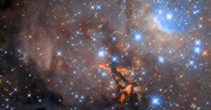 The field is filled with hundreds of bright stars. They are primarily blue in colour, with scattered smaller stars visible in yellow/orange. The background is dominated by cloudy grey dust, with permeating regions of dark black and orange.