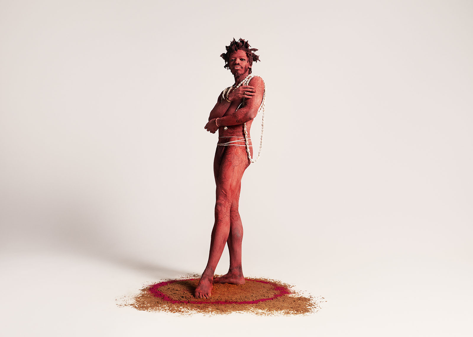 A man covered in red pigment stand above a pile of sand in the same color.