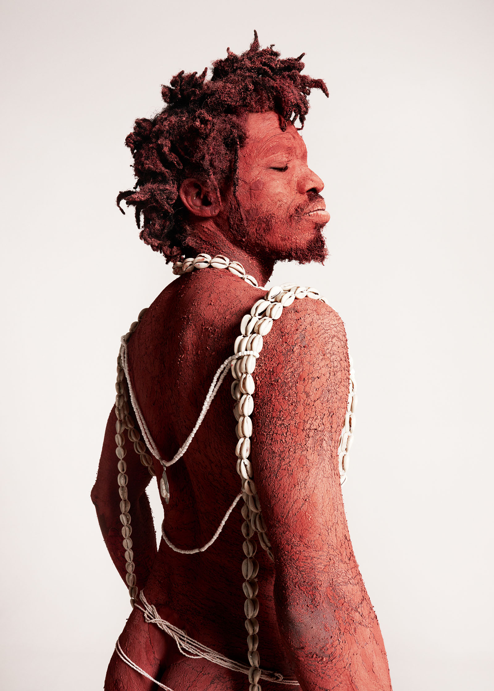 A man looks over his shoulder, covered in red pigment with shell necklaces draped over his back.
