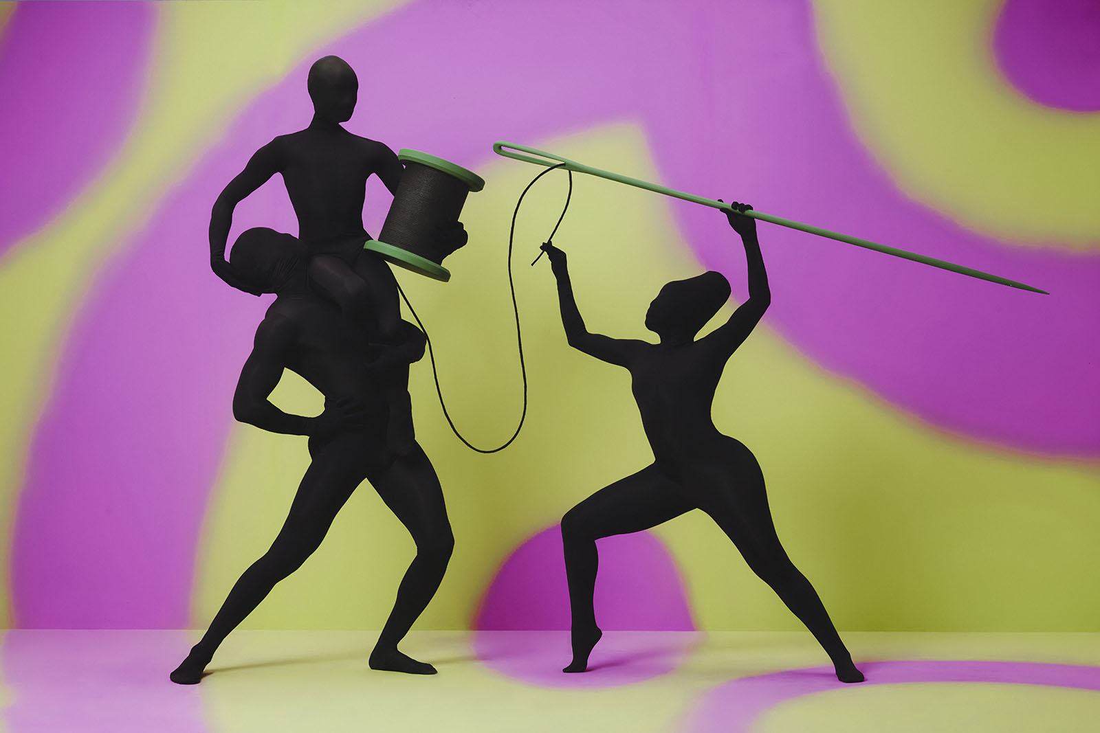 Three silhouettes pose holding a giant spool and giant sewing needle.