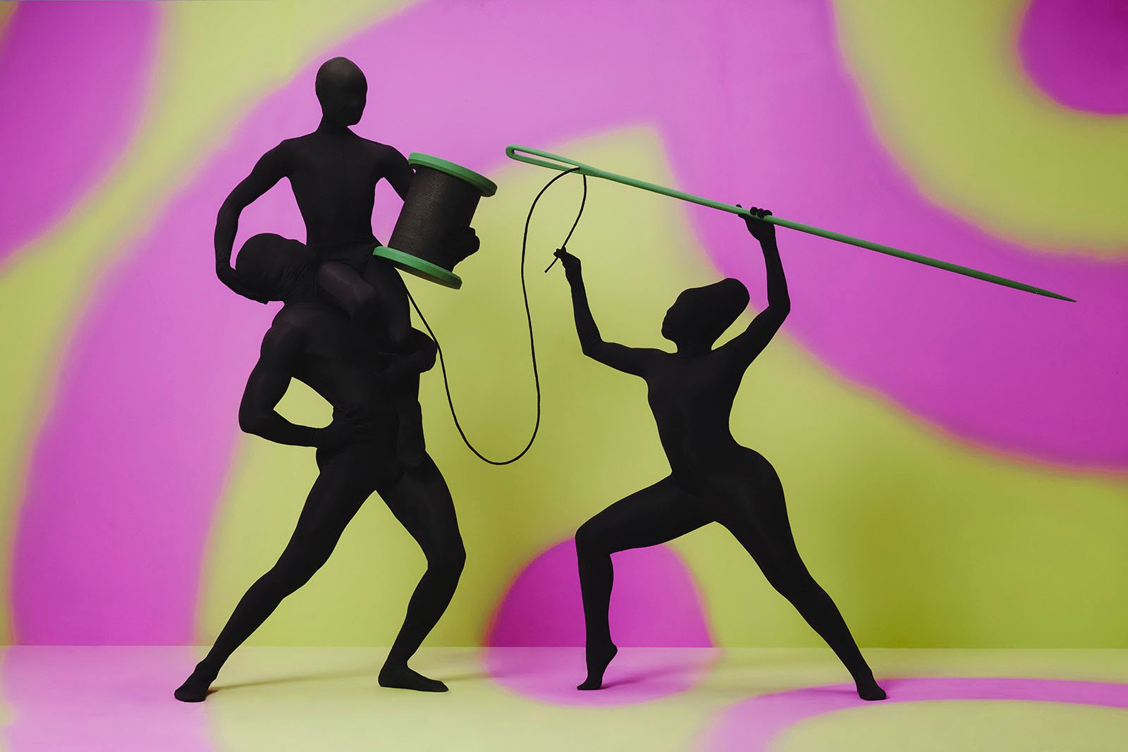 Three silhouettes pose holding a giant spool and giant sewing needle.