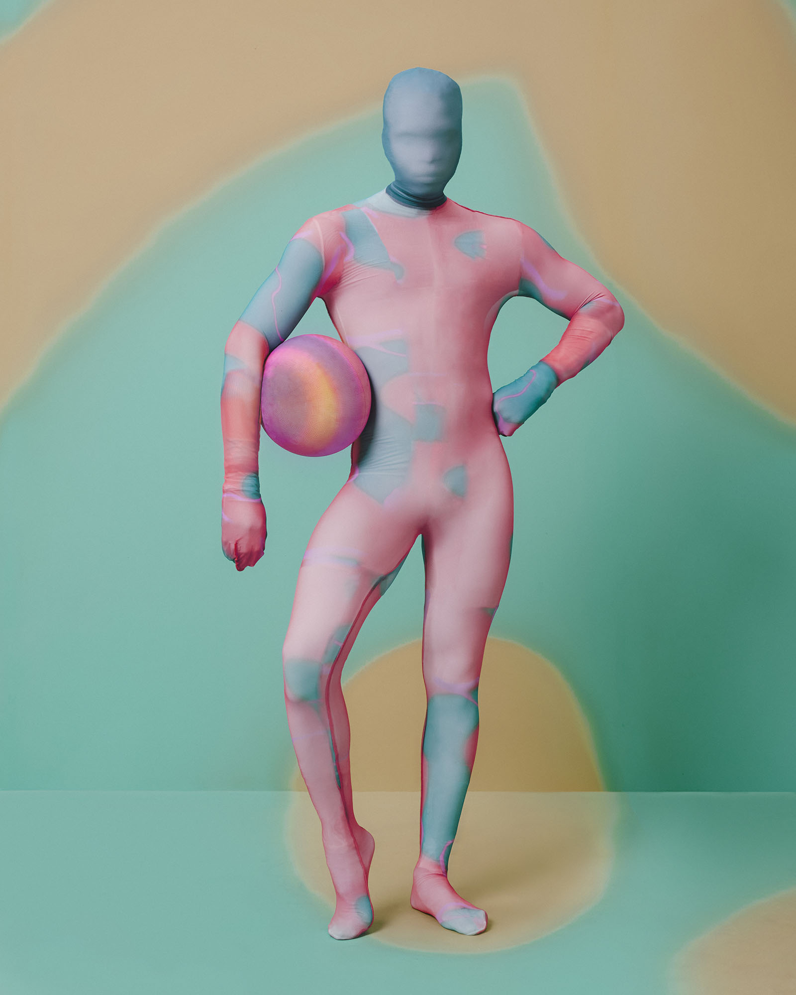 A person covered in a multicolored full-body suit holds a matching ball by their waist.