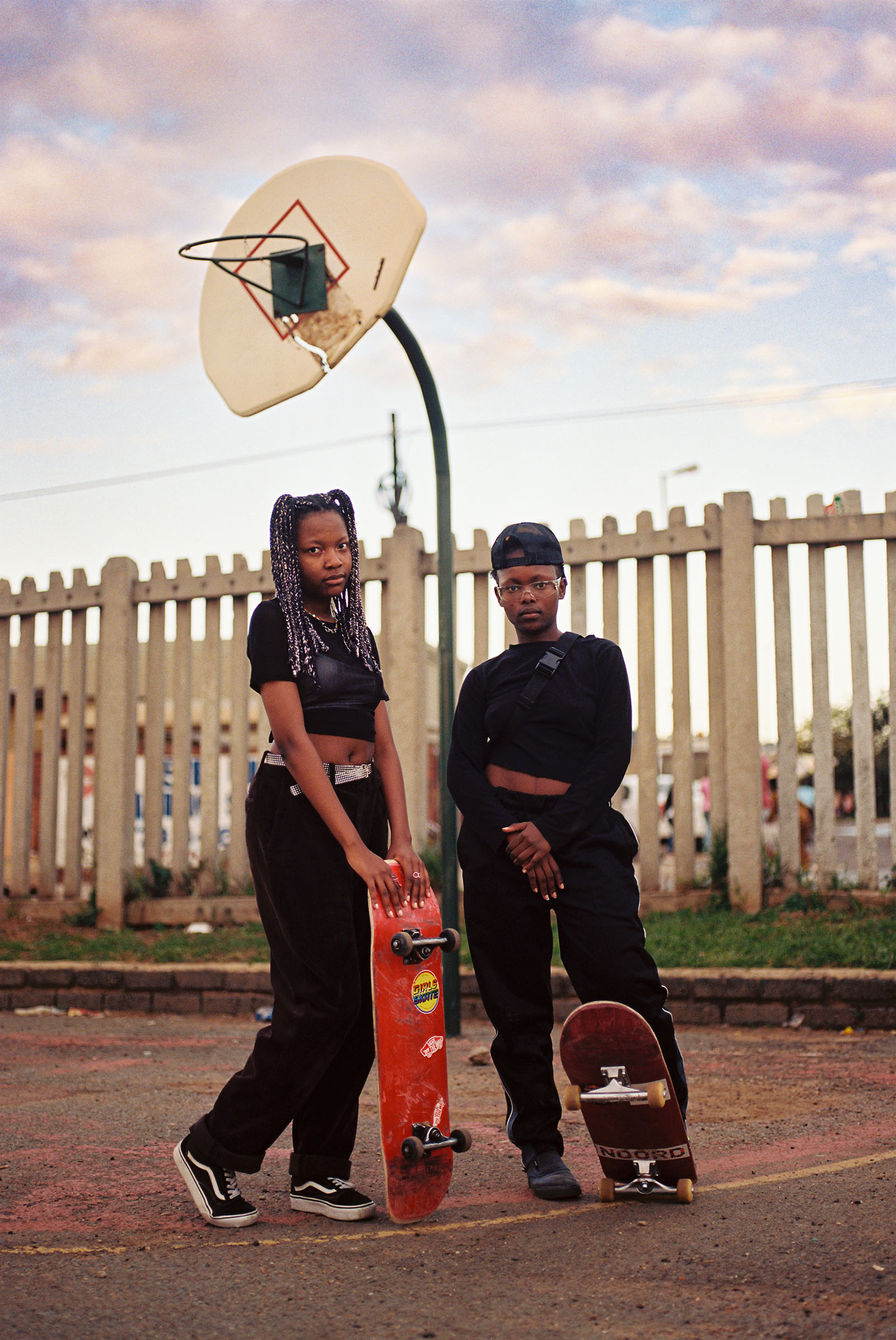 Two girls stand under a basketball hoop with skateboards.