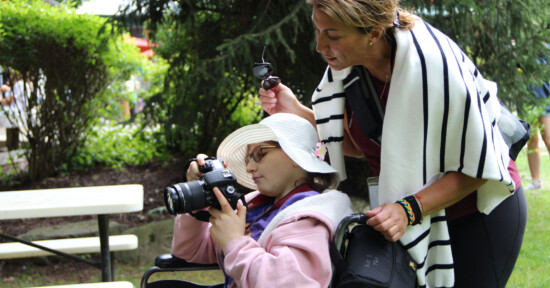 A child holds a Canon DSLR camera as she looks at an image she captured. A woman stands behind the child, looking at the image.