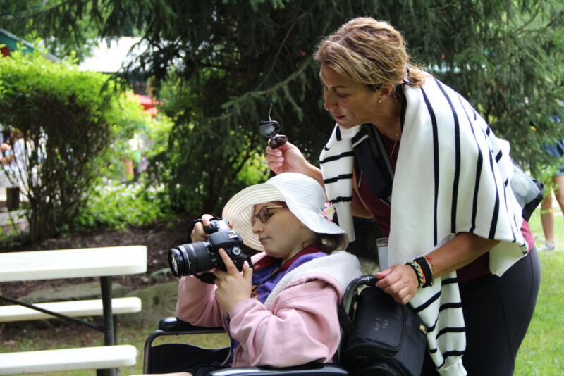 A child holds a Canon DSLR camera as she looks at an image she captured. A woman stands behind the child, looking at the image. 