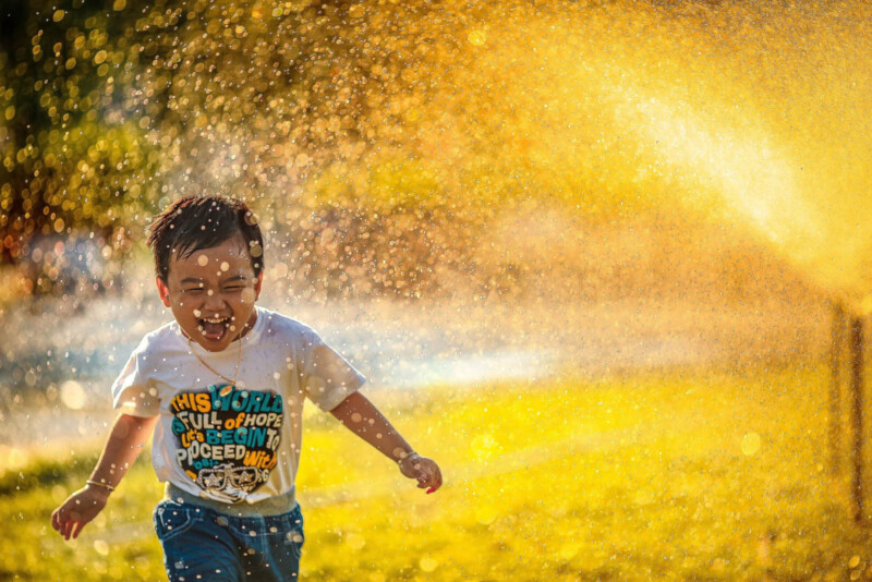 A young boy running through a water sprinkler with the sun in the background