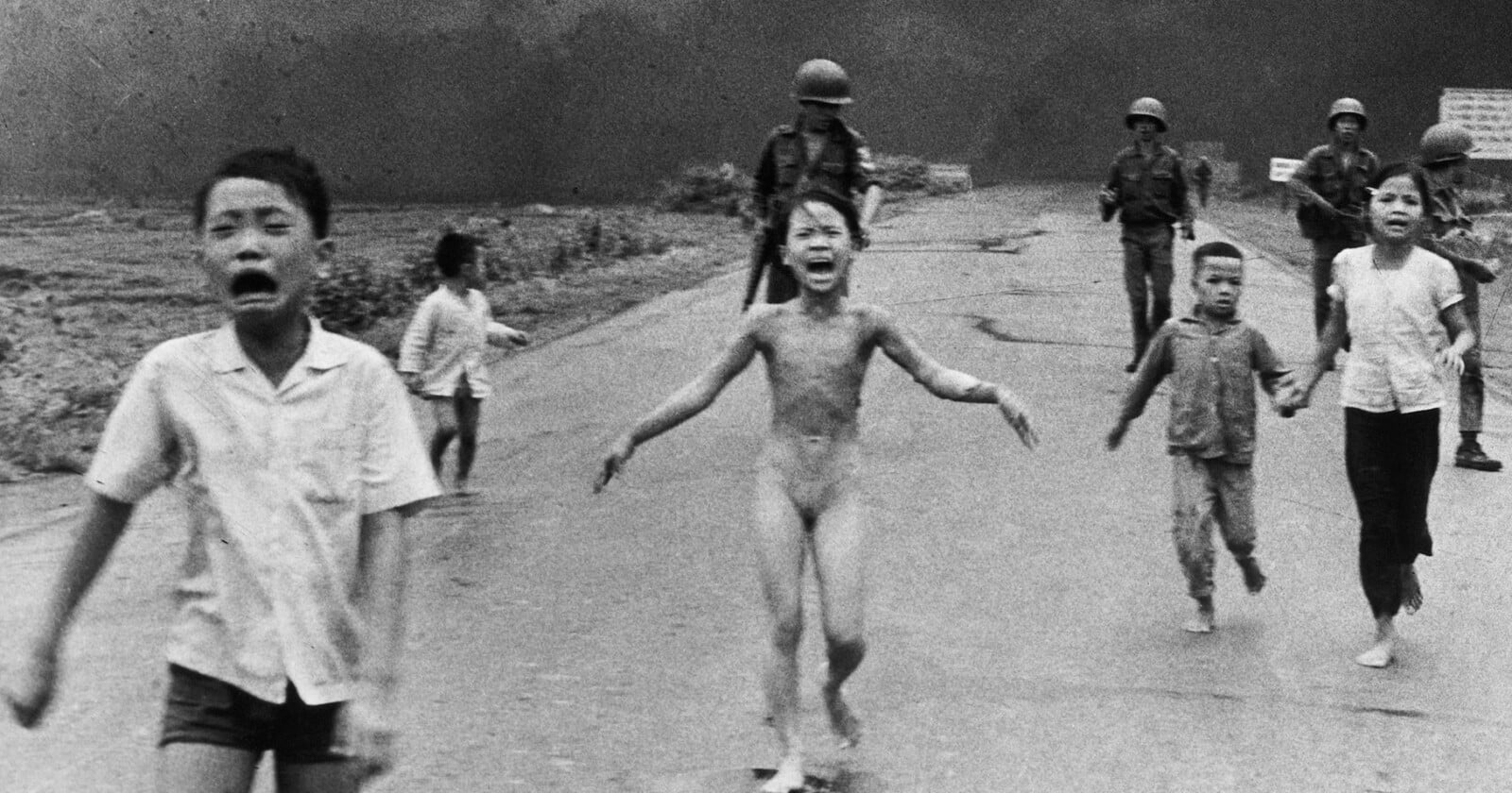 A group of children, some crying and barefoot, run down a road toward the camera with smoke and armed soldiers in the background. The central child appears distressed and unclothed.
