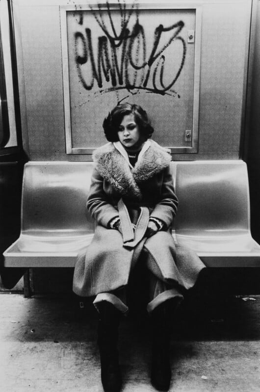 A woman sits on a subway underneath some graffiti.