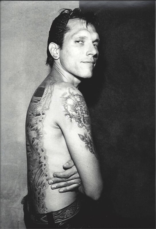 A shirtless man with tattoos looks back at the camera.