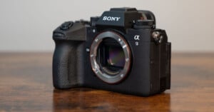 A sony α9 mirrorless camera with its lens removed, displaying its sensor, positioned on a wooden table against a blurred background.