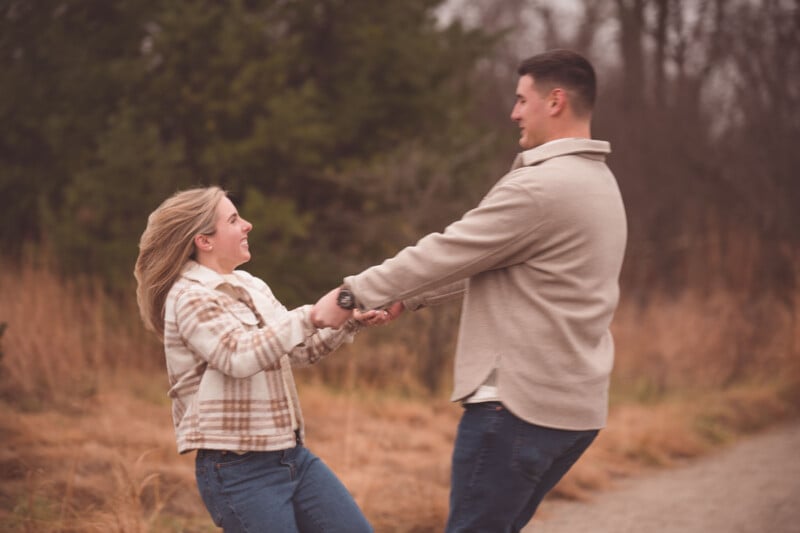 A couple holds hands spinning in a wooded area.