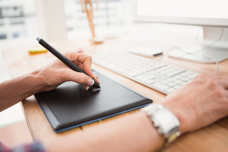 A hand holds the pen to an editing tablet while sitting in front a computer.