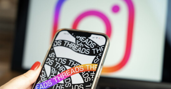 In a person's hand, a phone shows a screen from the Threads app in front of a computer displaying the Instagram logo.