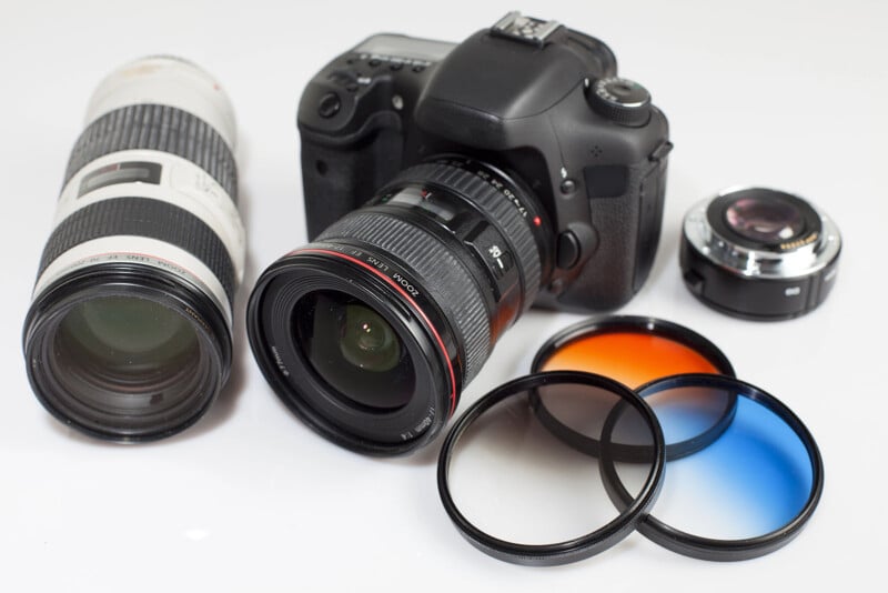 A camera sits on a white surface next to two lenses and three lens filters. 