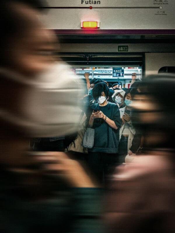 A person with a mask stands in front of a subway train with a crowd blurred while moving past. 