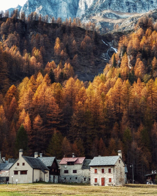 A small village sits in front of fall trees and a steep mountain cliff. 