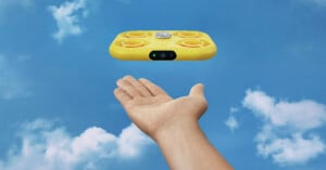 A yellow miniature drone hovers over a hand in front of a blue sky with a few clouds.