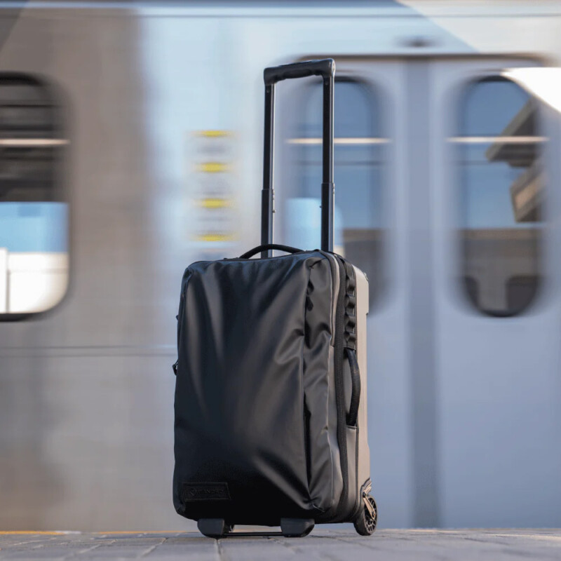 A Wandrd Tansit Carry-On Roller bag sits in front of a moving passenger train. 