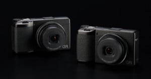 Two Ricoh GR series compact digital cameras are displayed against a black background. Both cameras have a sleek and minimalistic design, featuring prominent lenses and textured grips for easy handling. The branding "GR" is visible on the bottom right of each camera.