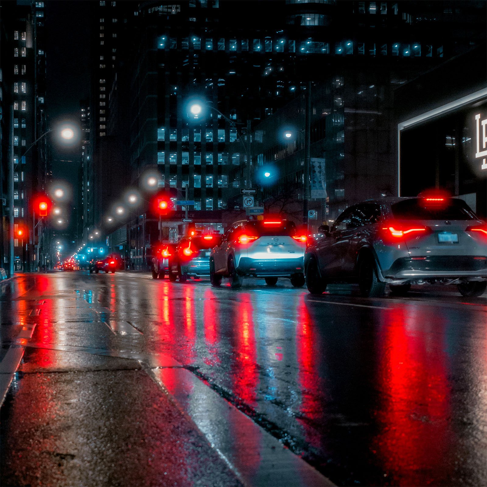 A city street at night is illuminated by streetlights and red taillights of cars lined up in traffic. The wet pavement reflects the vibrant lights, adding a colorful glow to the scene. Tall buildings with lit windows border the street.